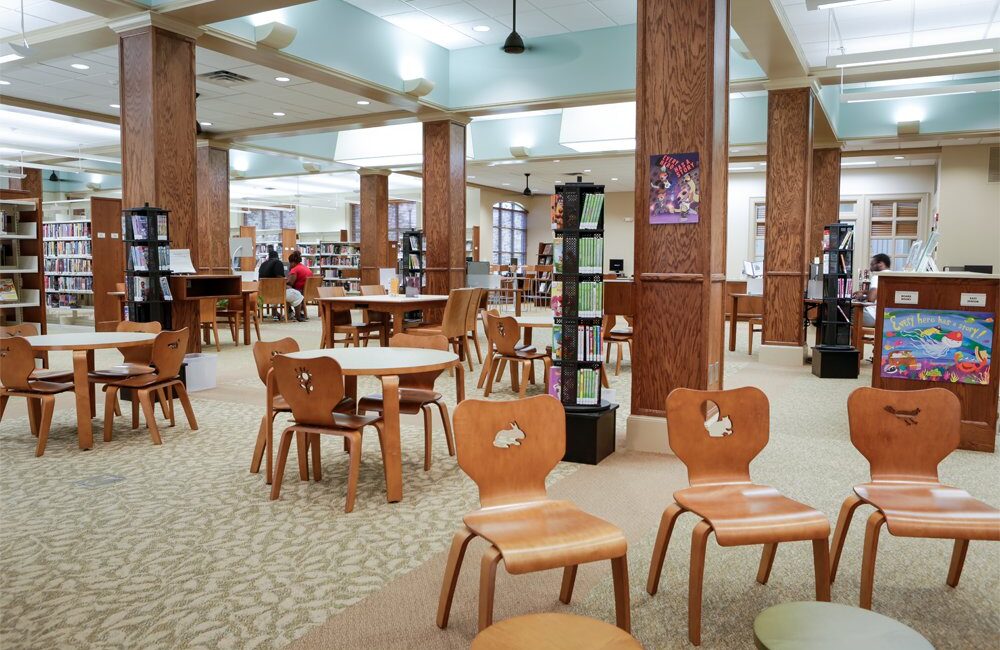 Spacious library children's area with wooden chairs, round tables, full bookshelves, colorful art, soft lighting, and people reading.