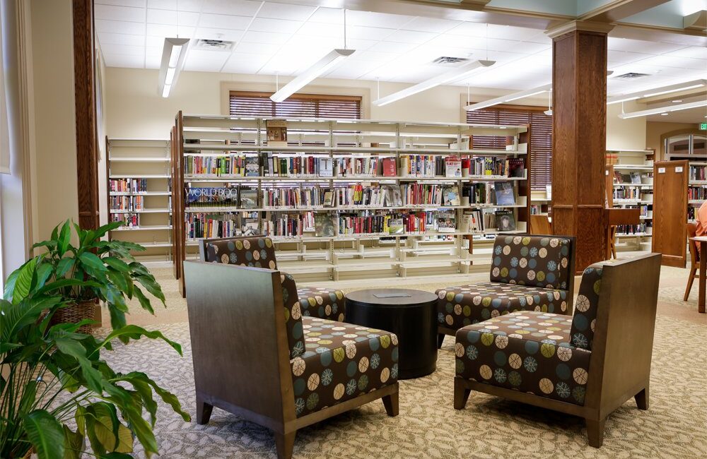 Cozy library seating with four patterned chairs around a round table on carpet. Book-filled shelves line the bright, well-lit space.