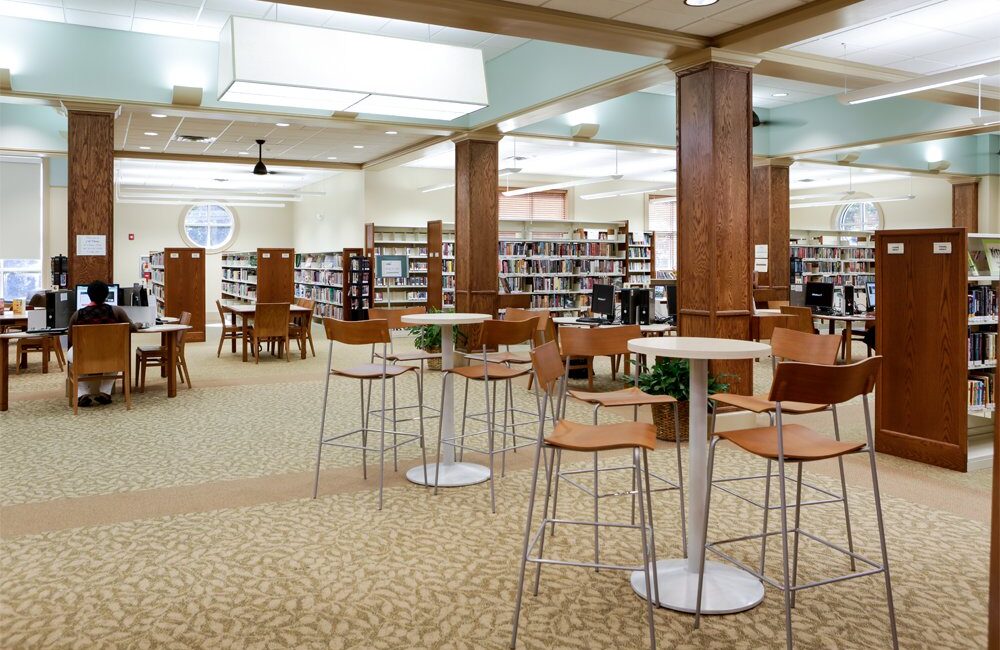 Spacious library with wooden bookshelves, high tables, computer stations, large sunlit windows, carpeted floors, and overhead lights.
