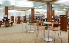 Spacious library with wooden bookshelves, high tables, computer stations, large sunlit windows, carpeted floors, and overhead lights.