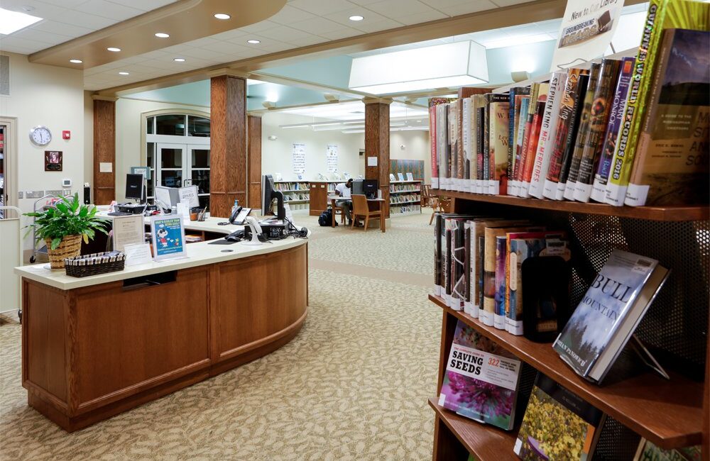Well-lit public library interior with bookshelves in front, a wooden desk with computers in the center, and more shelves and seating behind.