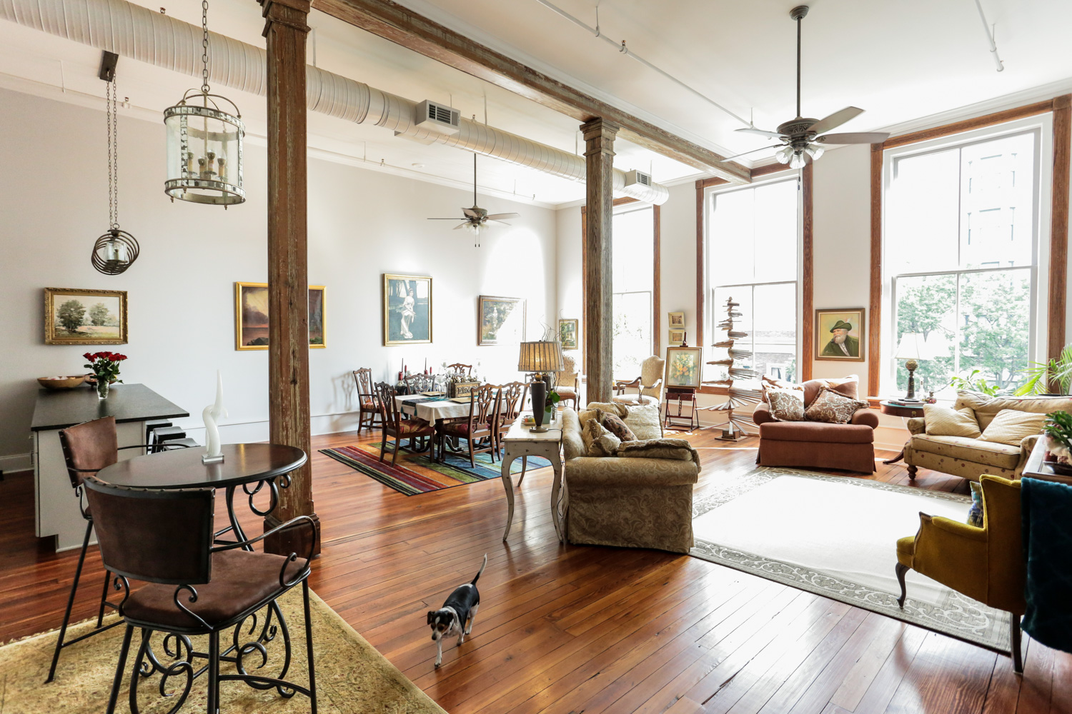 Bright living and dining area with big windows, wood floors, tall ceilings, vintage decor, wall art, and a small dog by a round table.