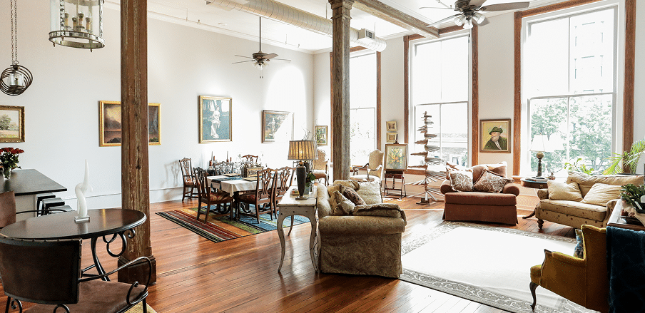 Spacious sunlit living and dining area with large windows, wood floors, vintage furniture, art on white walls, and tall columns.
