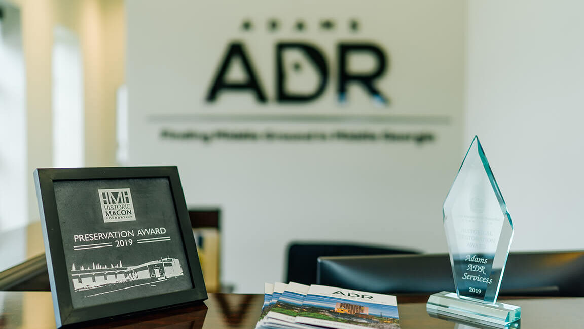 A framed 2019 Preservation Award, a crystal trophy, and brochures on a wooden desk with Adams ADR logo blurred in the background.