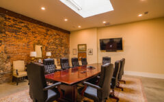 Conference room with a large wooden table, eight black leather chairs, brick accent wall, skylight, TV, framed art, and lamps.