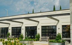 Modern building exterior with black-framed windows, beige brick walls, metal awnings, benches, planters, and a cloudy sky.