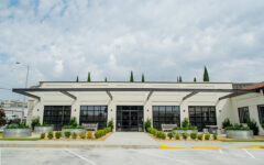 Modern single-story building with large black-framed windows, beige brick exterior, and landscaped entrance under partly cloudy sky.