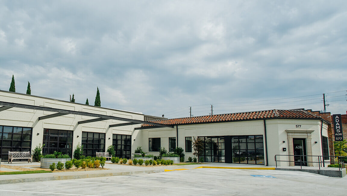 A modern white one-story commercial building with large windows, a tile roof, and ADAMS 577 sign by a mostly empty parking lot under clouds.