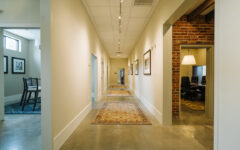 Bright hallway with polished concrete floors, beige walls, rugs, framed art, and open doors to modern and rustic rooms with brick accents.