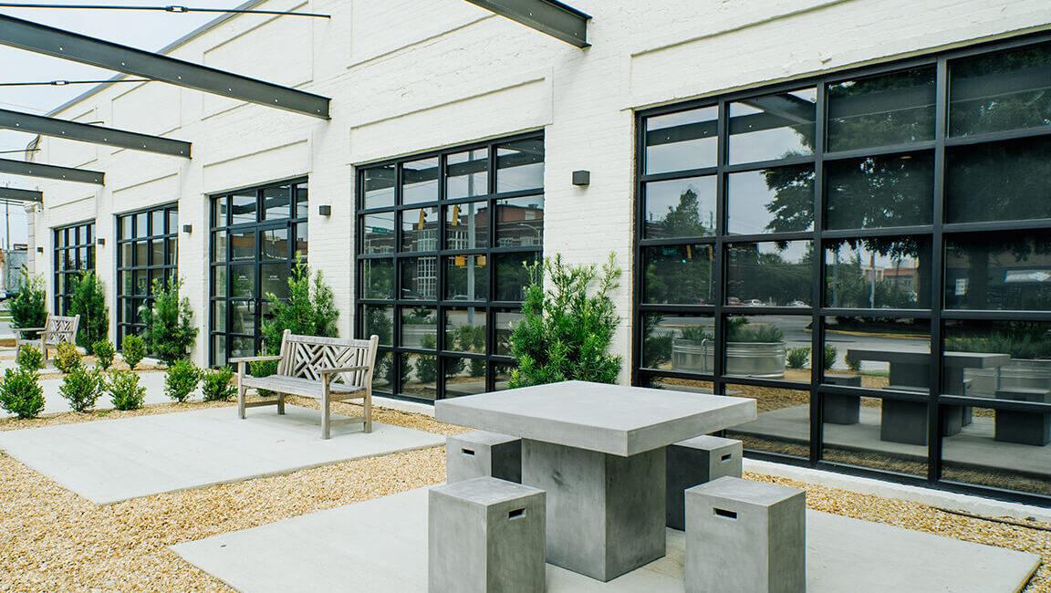 Modern outdoor patio with concrete table, stools, and wooden bench on gravel next to a white building with black-framed windows.