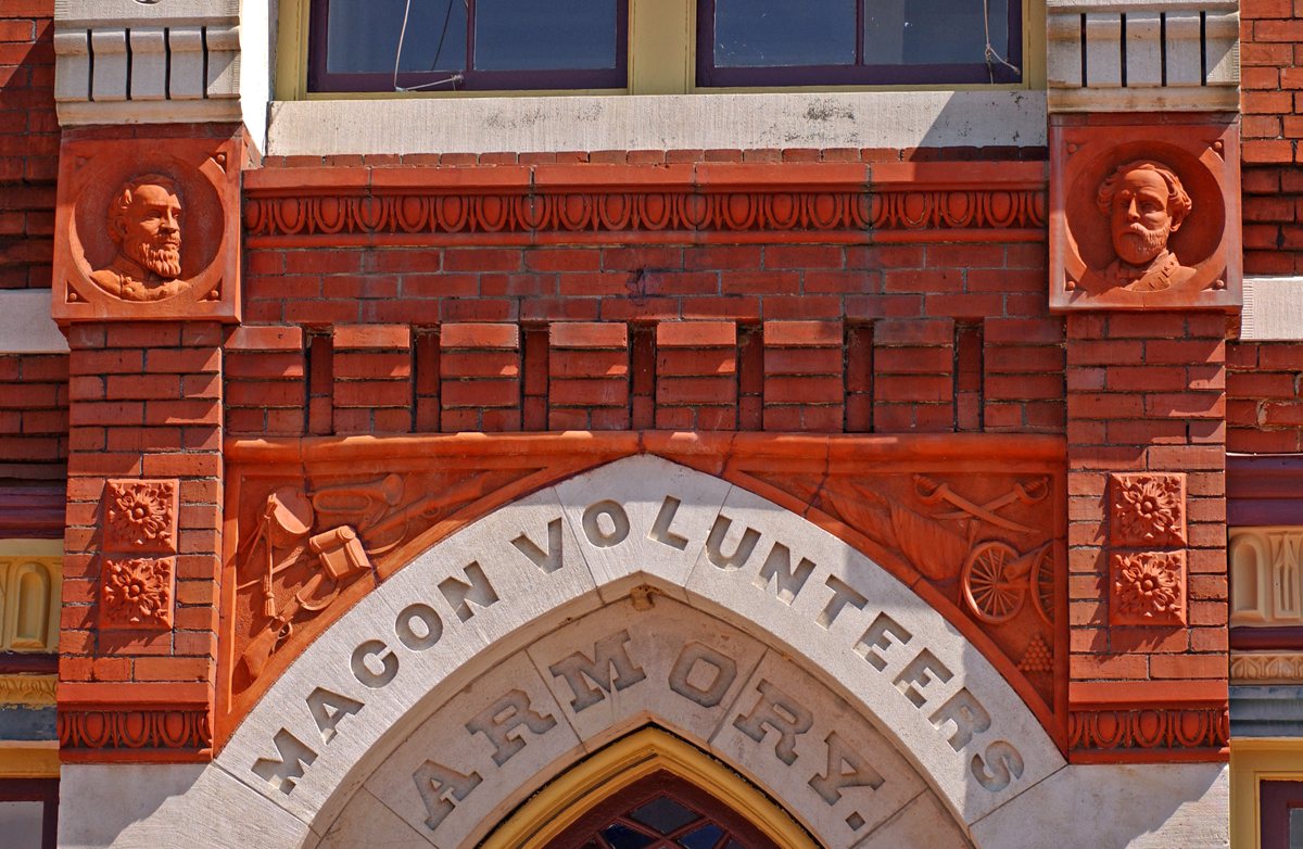 A brick building facade with MACON VOLUNTEERS ARMORY carved above an arched entrance, reliefs of bearded men, and brick symbols.