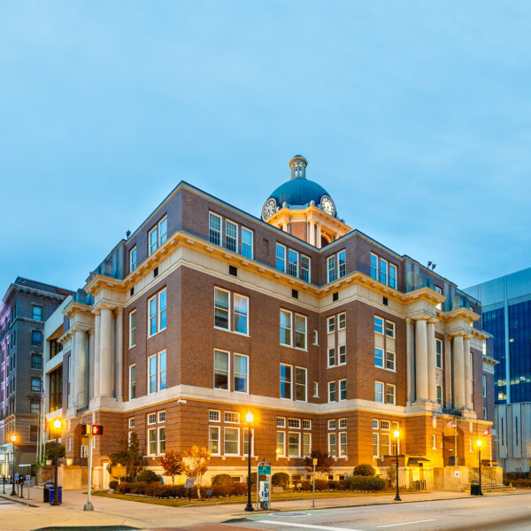 A grand historic brick building with a green-domed clock tower and columns, illuminated by streetlights at dusk on a city corner.