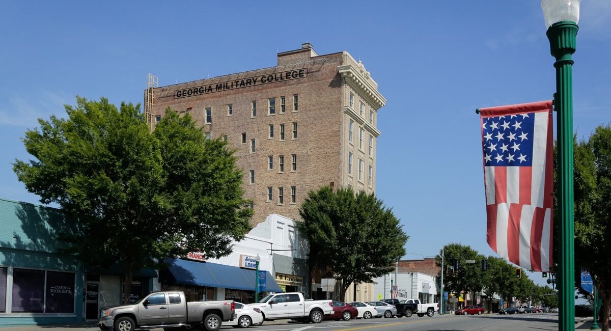 A street view in a small town with parked cars, storefronts, Georgia Military College building, flag banner, trees, blue sky.