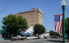 A street view in a small town with parked cars, storefronts, Georgia Military College building, flag banner, trees, blue sky.