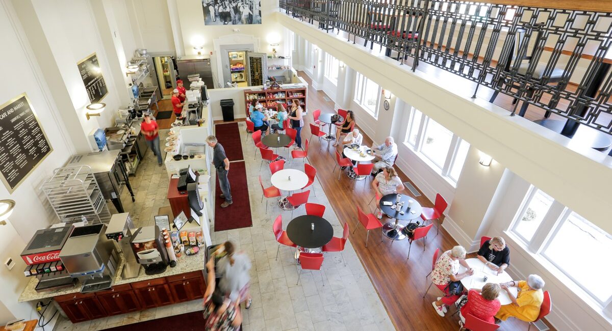 Bright, airy café with red chairs and white tables. Guests dine and chat while staff work behind the counter. Large windows, balcony above.