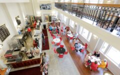 Bright, airy café with red chairs and white tables. Guests dine and chat while staff work behind the counter. Large windows, balcony above.