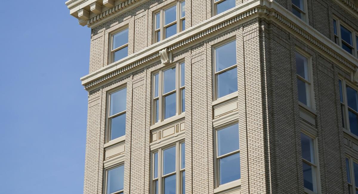 A beige brick building with decorative cornices and large windows stands under a blue sky, topped with ornate architectural moldings.