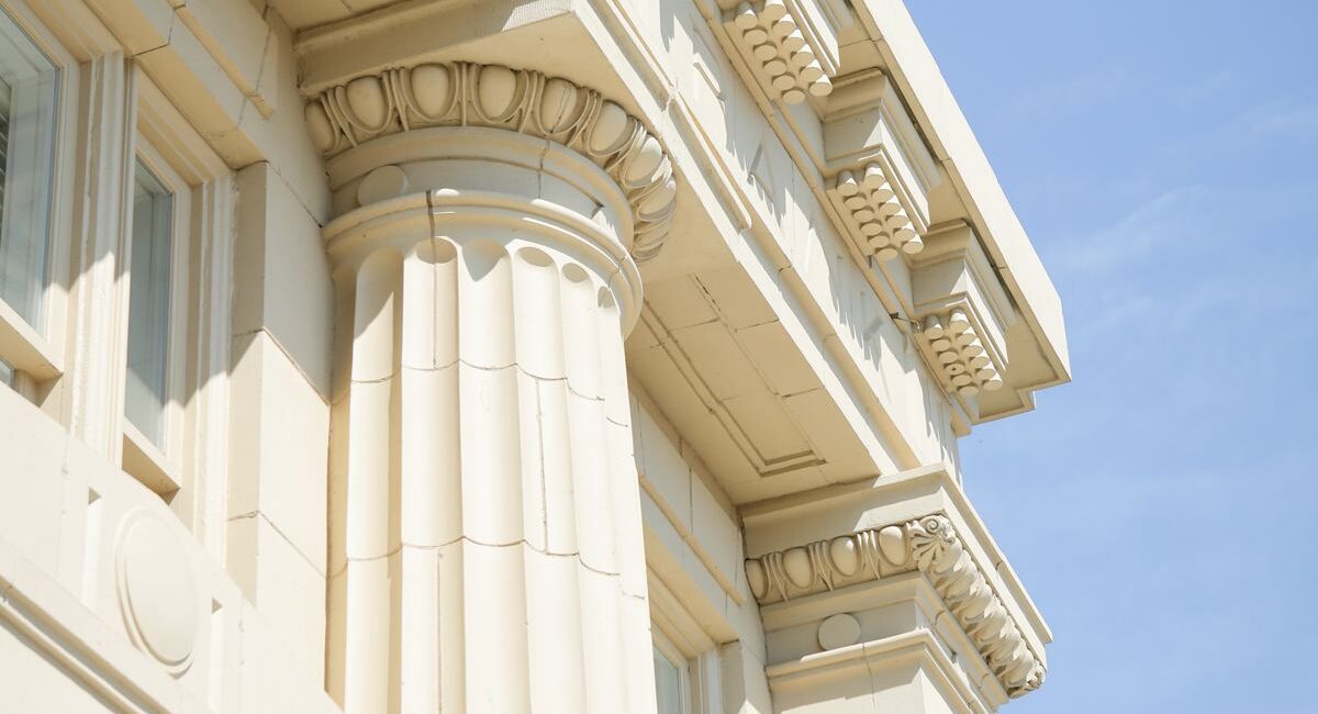 Close-up view of a beige stone building with fluted columns, ornate cornices, and classical details under a clear blue sky.