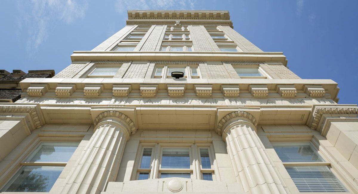 Low-angle shot of a historic beige stone building, tall columns and large windows, ornate details, under a clear blue sky.