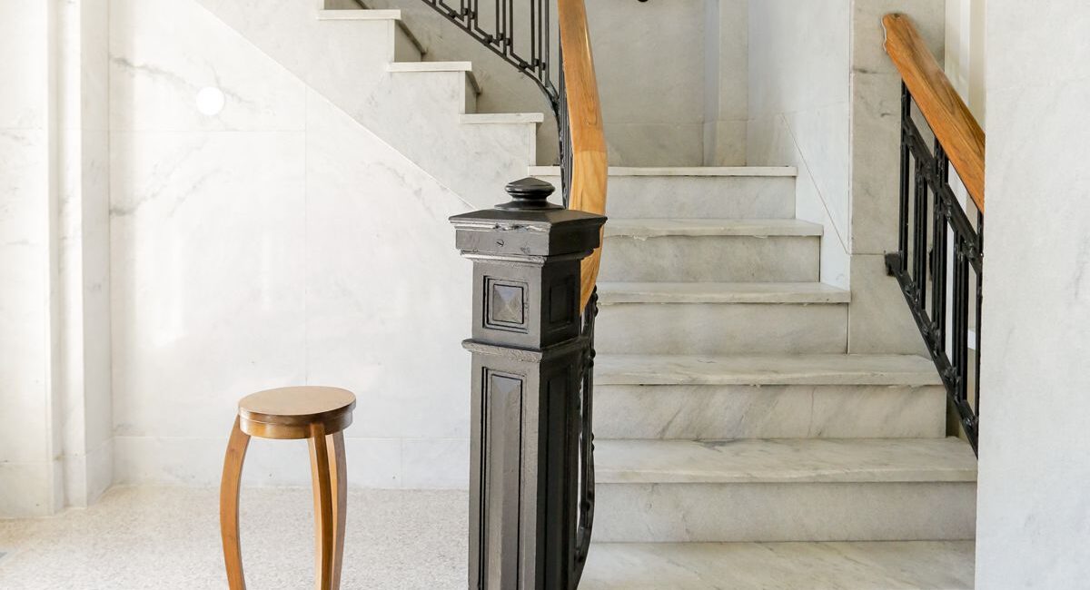Marble stairs with black and wood railings curve upward in a sunny, bright space. A round wooden side table stands at the base.