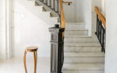 Marble stairs with black and wood railings curve upward in a sunny, bright space. A round wooden side table stands at the base.