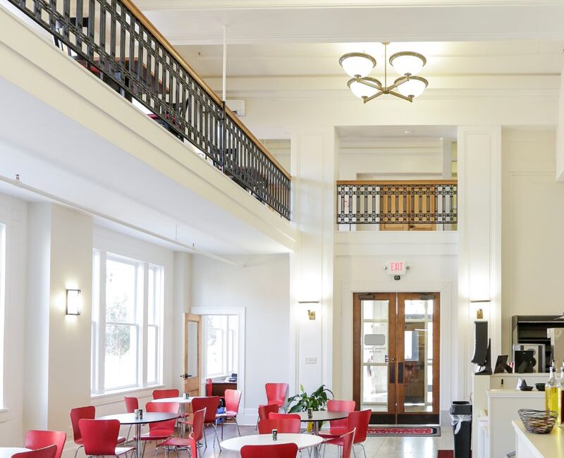 A bright modern café with red chairs, round tables, marble floors, a red rug, white walls, large windows, and chandeliers above.