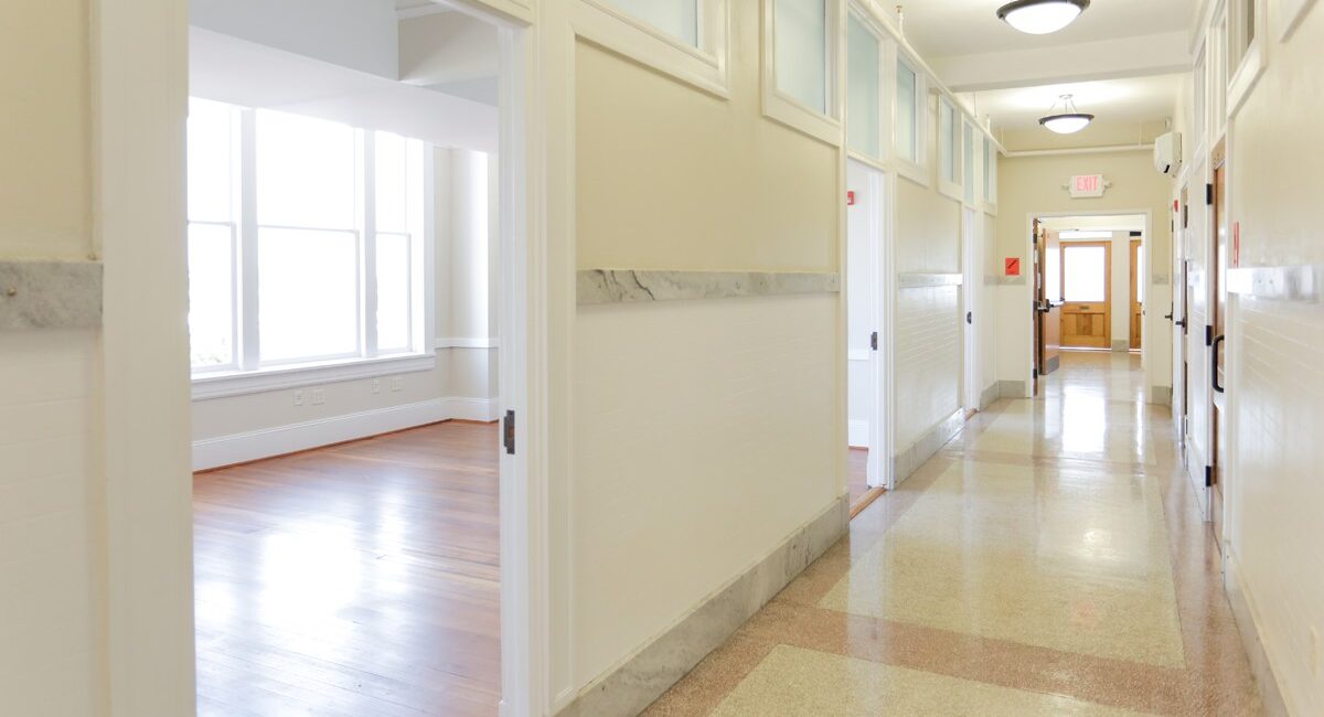 Bright, empty hallway with cream walls, terrazzo floors, ceiling lights, and doorway leading to sunny room with large windows and wood floors.