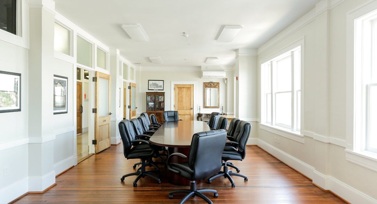 Bright conference room with a long oval table, black chairs, large windows, wooden floors, and framed pictures on white walls.