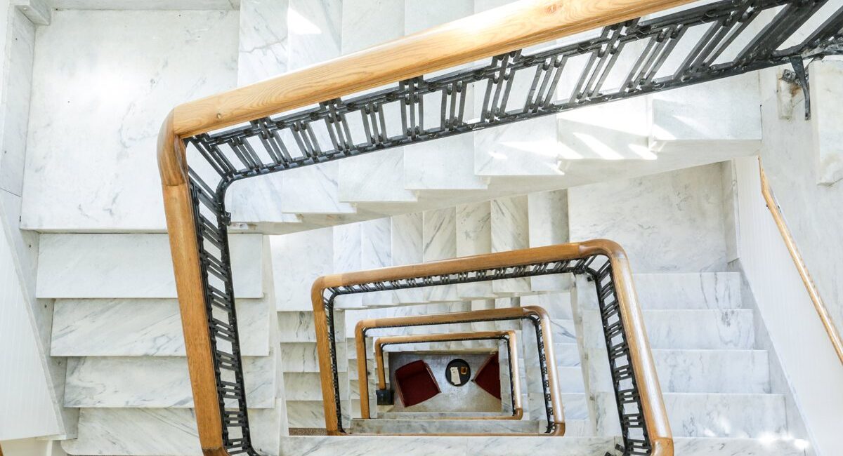 Top-down view of a white marble spiral staircase with wooden railings, black balusters, red chairs, and a small table below.