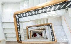 Top-down view of a white marble spiral staircase with wooden railings, black balusters, red chairs, and a small table below.