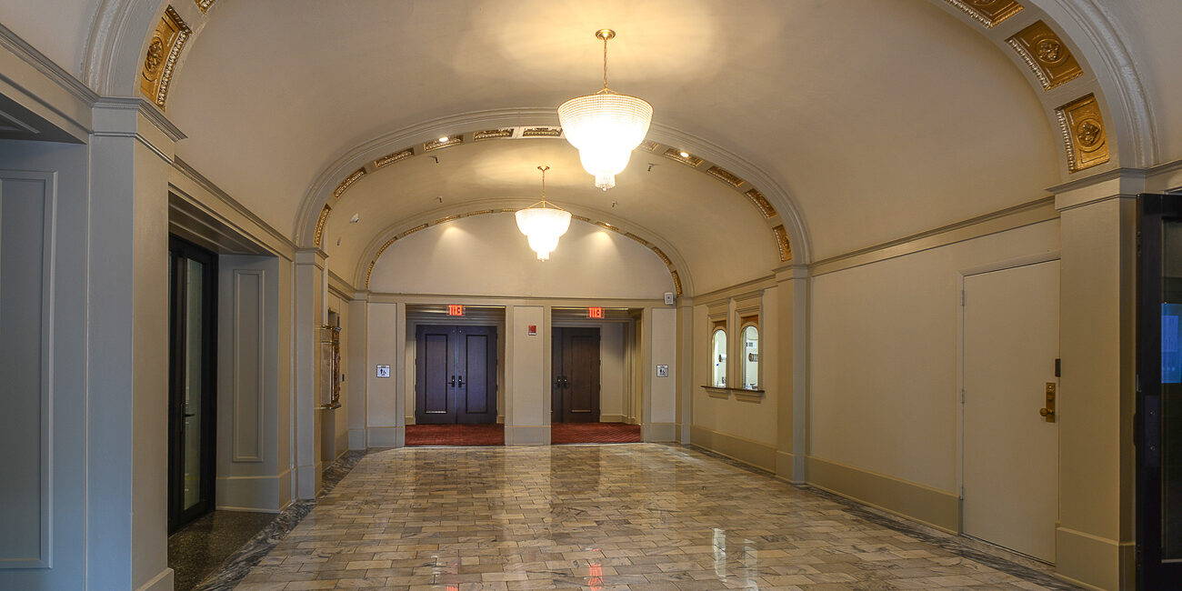 A hallway with polished marble floors, a gold-accented arched ceiling, ornate chandeliers, elevator doors at the end, and double doors on both sides.