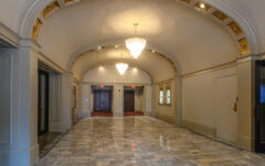 A hallway with polished marble floors, a gold-accented arched ceiling, ornate chandeliers, elevator doors at the end, and double doors on both sides.