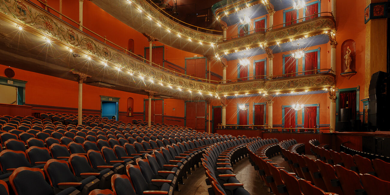 Elegant empty theater with rows of blue seats, ornate balconies, warm lighting, decorative railings, and red curtains throughout.