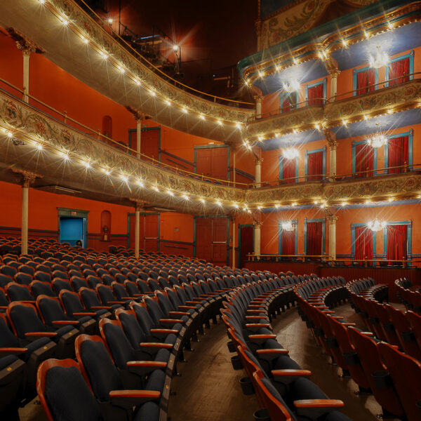 Elegant empty theater with rows of blue seats, ornate balconies, warm lighting, decorative railings, and red curtains throughout.