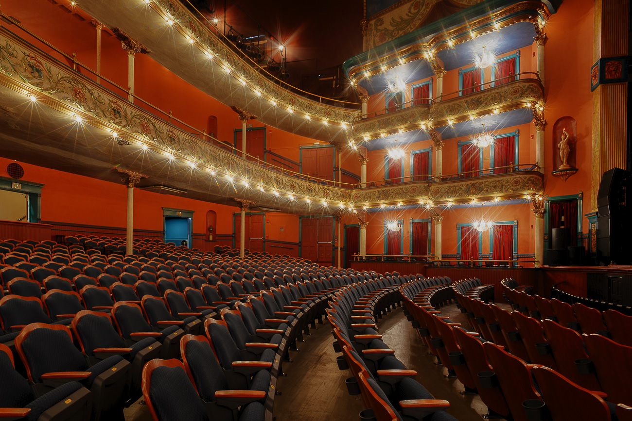 Elegant empty theater with rows of blue seats, ornate balconies, warm lighting, decorative railings, and red curtains throughout.