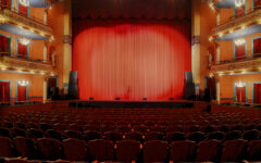 Empty red theater seats face a stage with a closed red curtain, ornate balconies, gold accents, and glowing wall sconces.