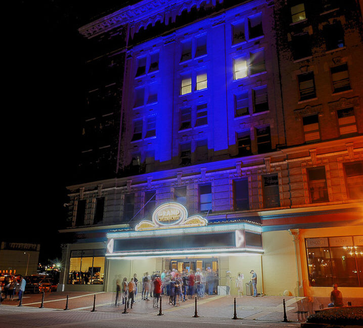 A group of people stand outside a theater entrance at night, marquee glowing; the building is lit with blue and orange lighting.