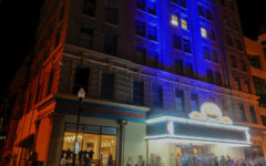 A large theater at night shines with blue and purple lights as people enter and pass by, with a street lamp lighting the sidewalk.