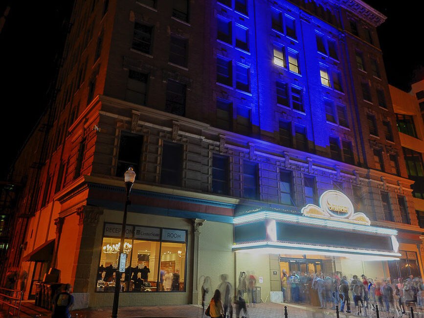 A large theater at night shines with blue and purple lights as people enter and pass by, with a street lamp lighting the sidewalk.