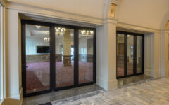 Glass sliding doors divide a marble hallway from a carpeted room with chandeliers; gold trim and arches adorn the hallway ceiling.
