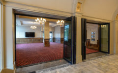 Large elegant room with open double doors, red patterned carpet, chandeliers, wood paneling, marble tile floor, and TV in back.