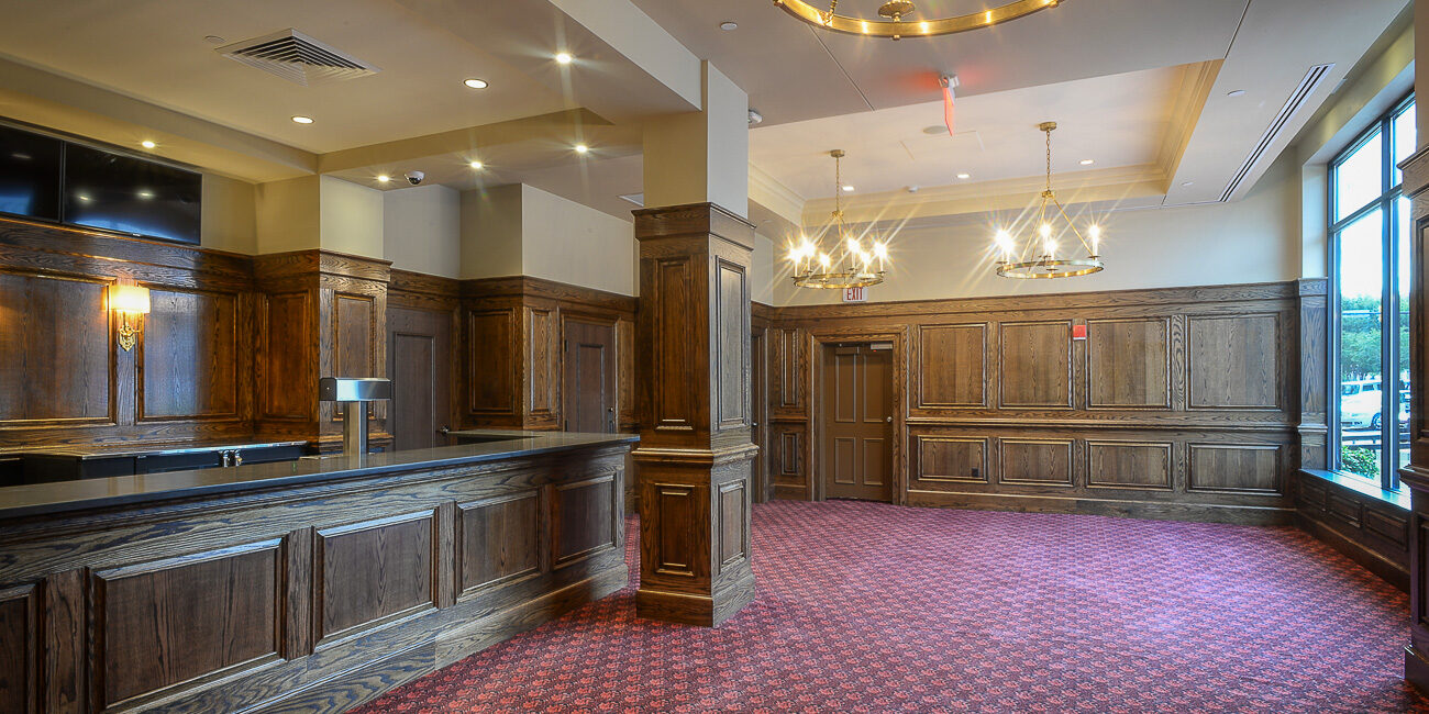 Elegant room with dark wood paneling, wooden counter, red patterned carpet, large windows, and two gold chandeliers overhead.