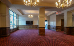 A spacious empty room with red patterned carpet, beige walls, wood paneling, large windows, chandeliers, and a wall-mounted TV.