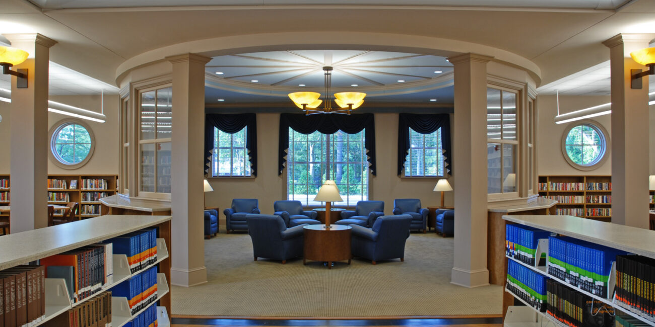 Spacious, bright library with bookshelves, circular windows, blue armchairs around a table, pendant lights, and tall dark-draped windows.