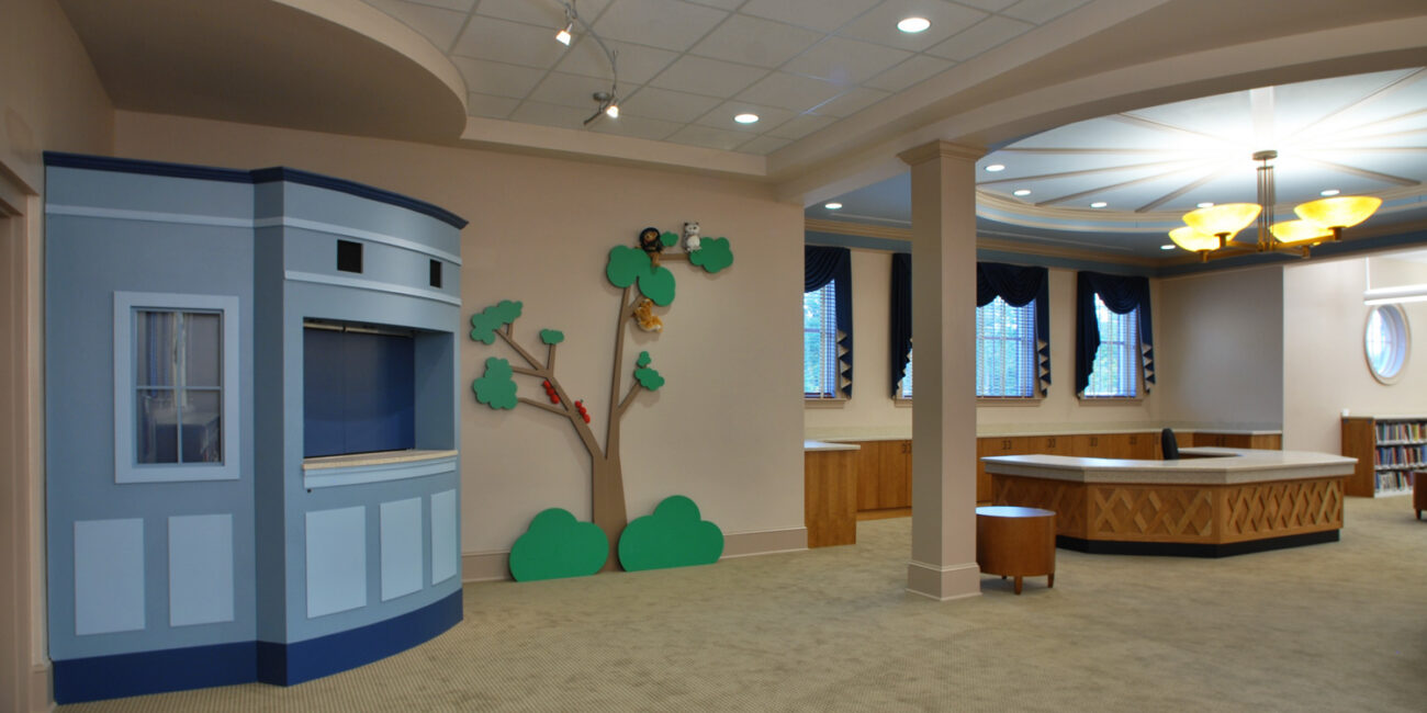A bright, roomy library features a blue puppet theater, wall art of a tree, big windows with dark curtains, light wood tables, and beige carpet.