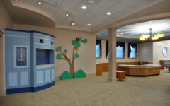A bright, roomy library features a blue puppet theater, wall art of a tree, big windows with dark curtains, light wood tables, and beige carpet.