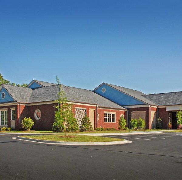A large red-brick building with a gray roof and white trim sits by an empty parking lot, surrounded by trees under a clear blue sky.