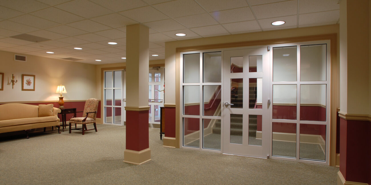 A spacious carpeted lobby with beige walls, sofa, armchair, lamp, framed art on the left, and glass doors to a stairwell.