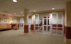 A spacious carpeted lobby with beige walls, sofa, armchair, lamp, framed art on the left, and glass doors to a stairwell.