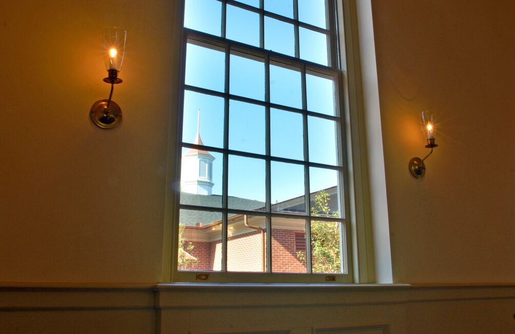 Tall arched church window with sunlight streaming in, steeple visible outside, and lit sconces mounted on the interior wall.